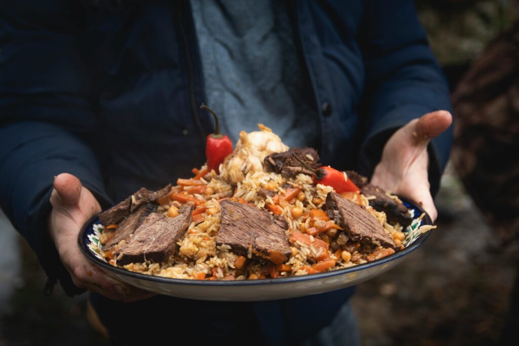 Close-up of a person holding a plate of beef pilaf with vegetables outdoors.