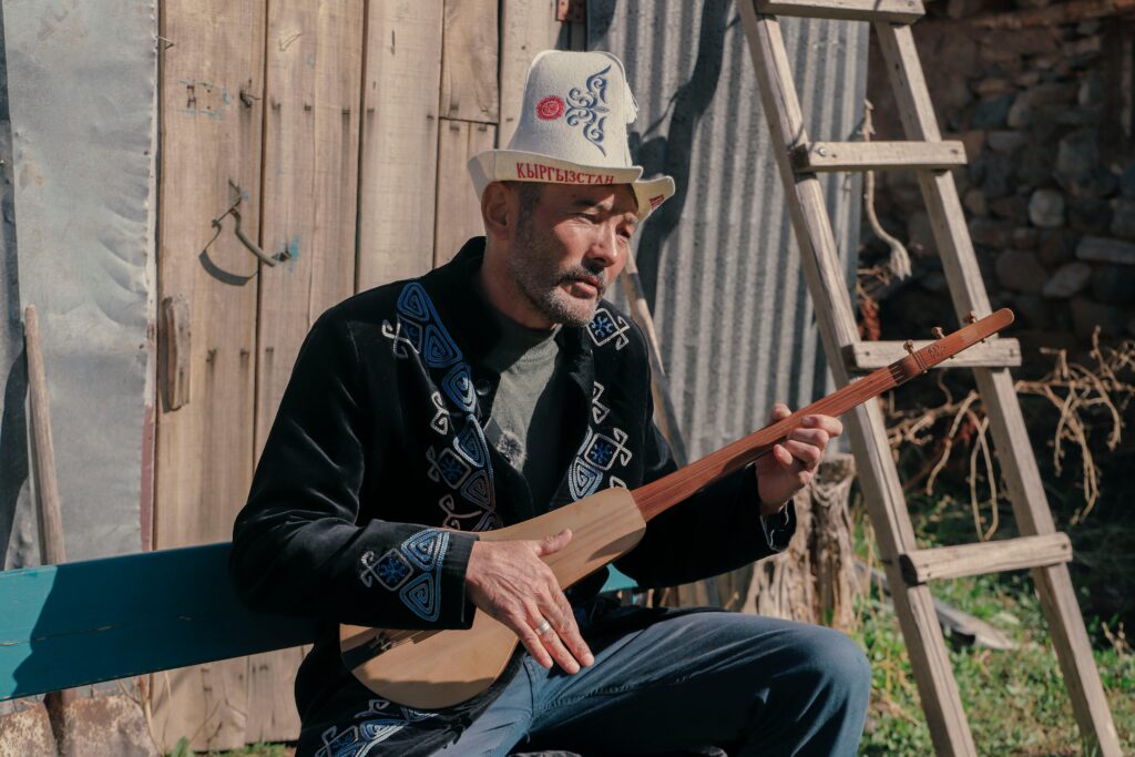 A musician from Kyrgyzstan plays a traditional string instrument outdoors, wearing cultural attire.