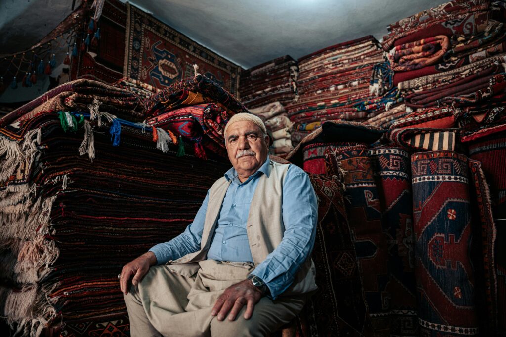 An elderly man sits surrounded by traditional handmade carpets in a shop.