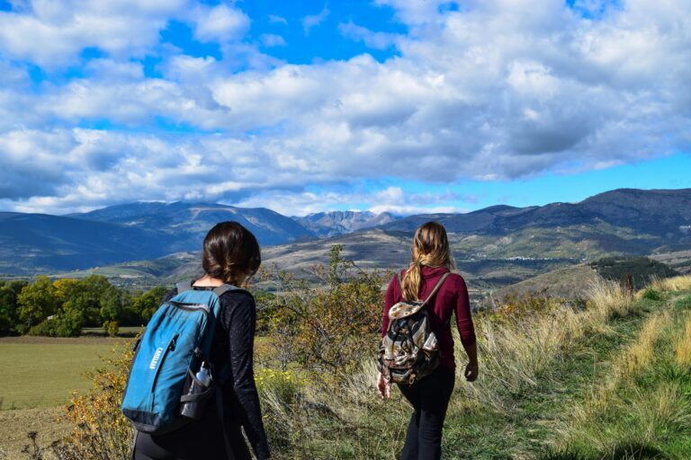 Deux femmes faisant de la randonnée sur un sentier de montagne avec des vues panoramiques sous un ciel bleu.