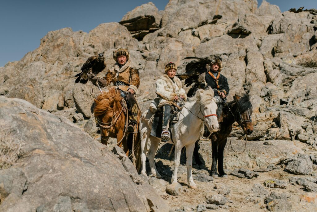 Focused kyrgyz males in traditional warm wear riding horses and carrying eagles during eagle hunting at mountainous terrain