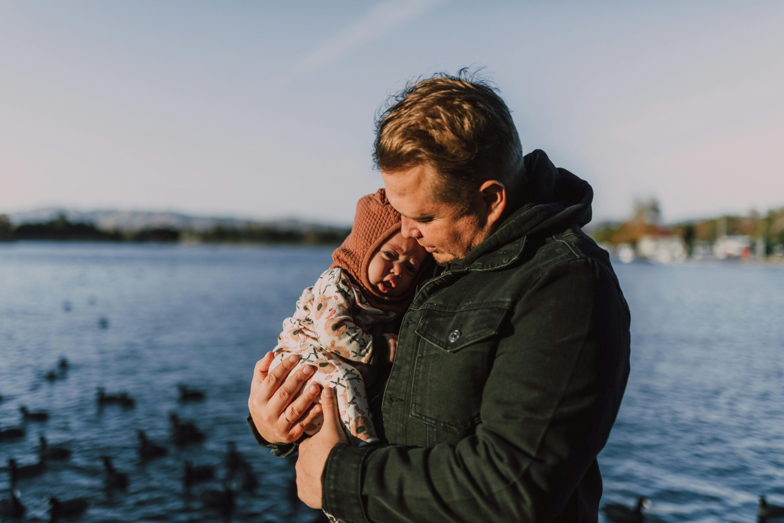 A heartwarming portrait of a father holding his baby by the lakeside, surrounded by nature.