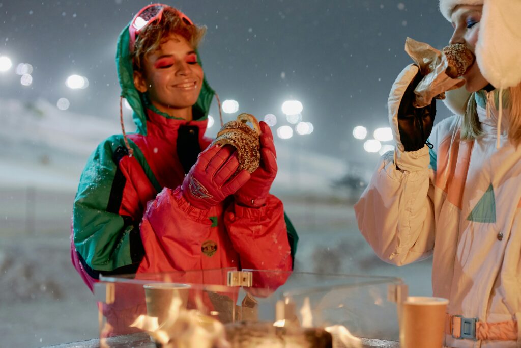 Two women enjoy hot pastries near a fire after snowboarding at night.