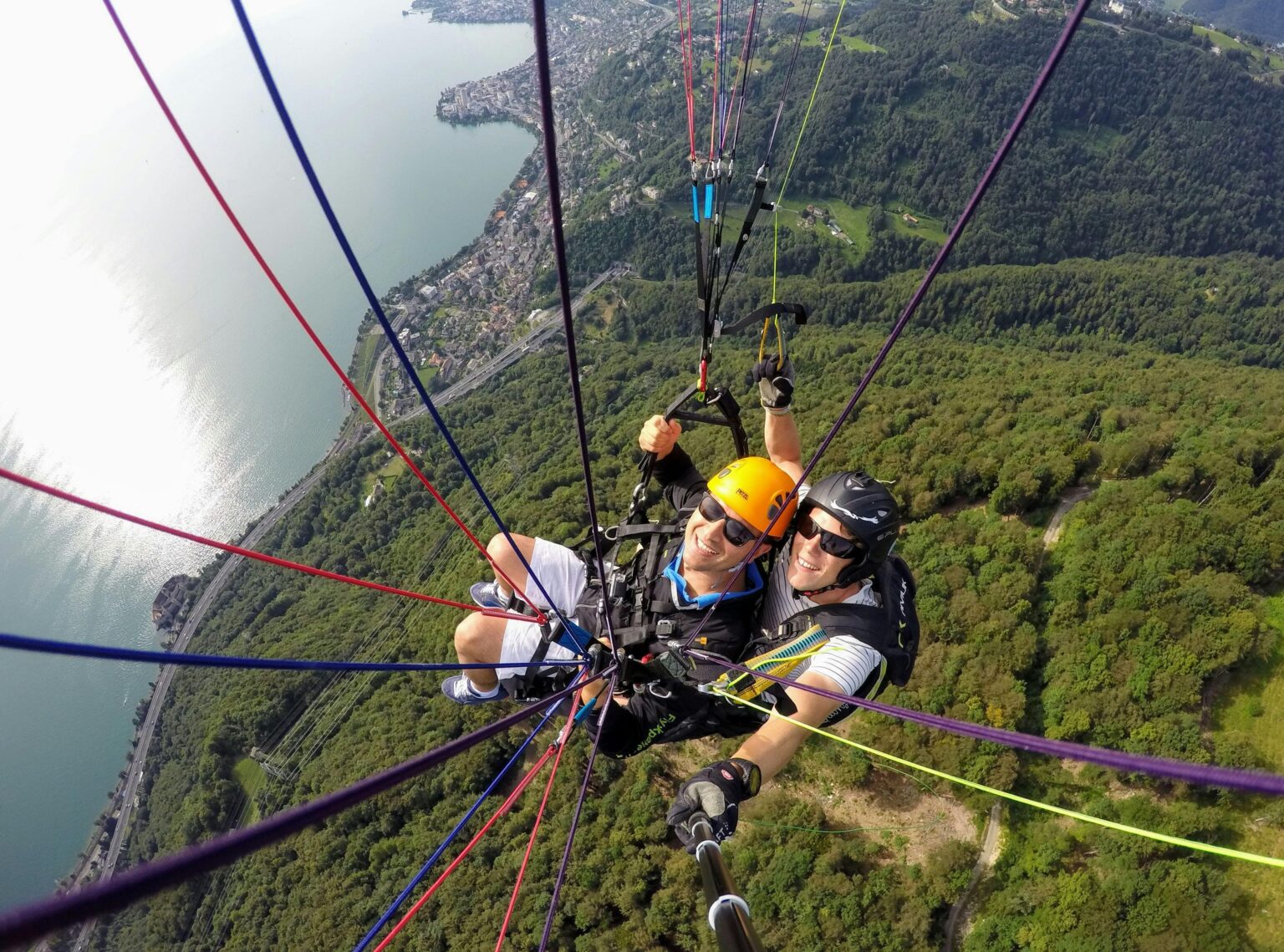 Two adults paragliding over a scenic landscape with forest and lake views, captured from above.
