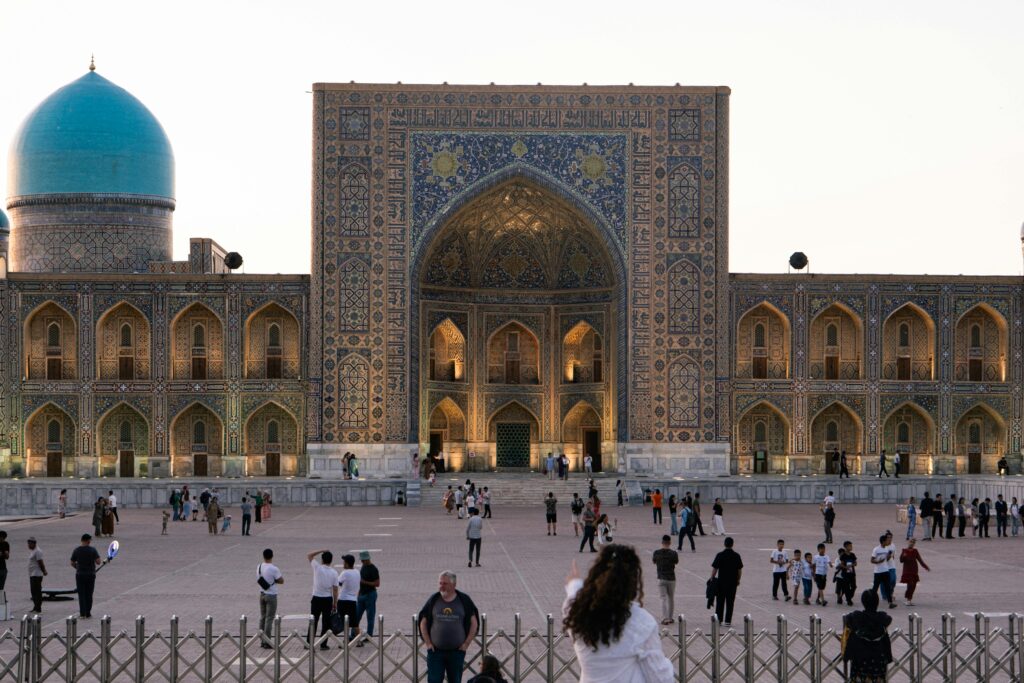 People gathering at the illuminated Registan complex in Samarkand at dusk.