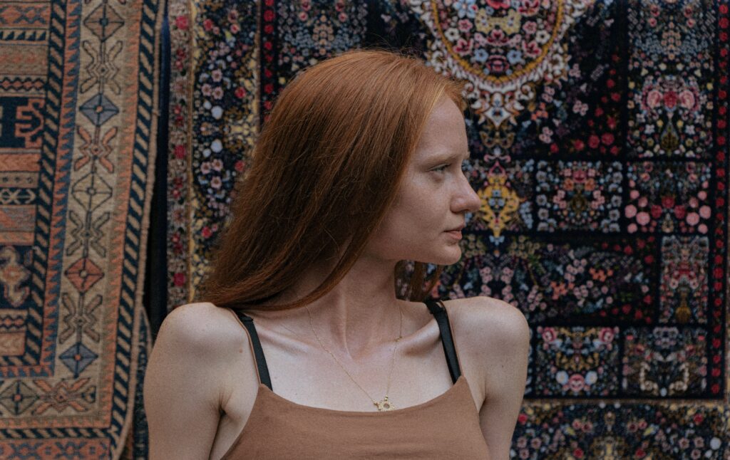 A woman with red hair stands in front of ornate rugs, showcasing cultural patterns in Baku, Azerbaijan.