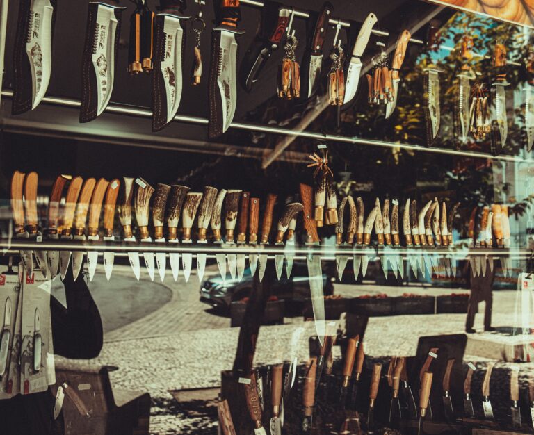 A variety of knives displayed in a shop window reflecting city street scene.